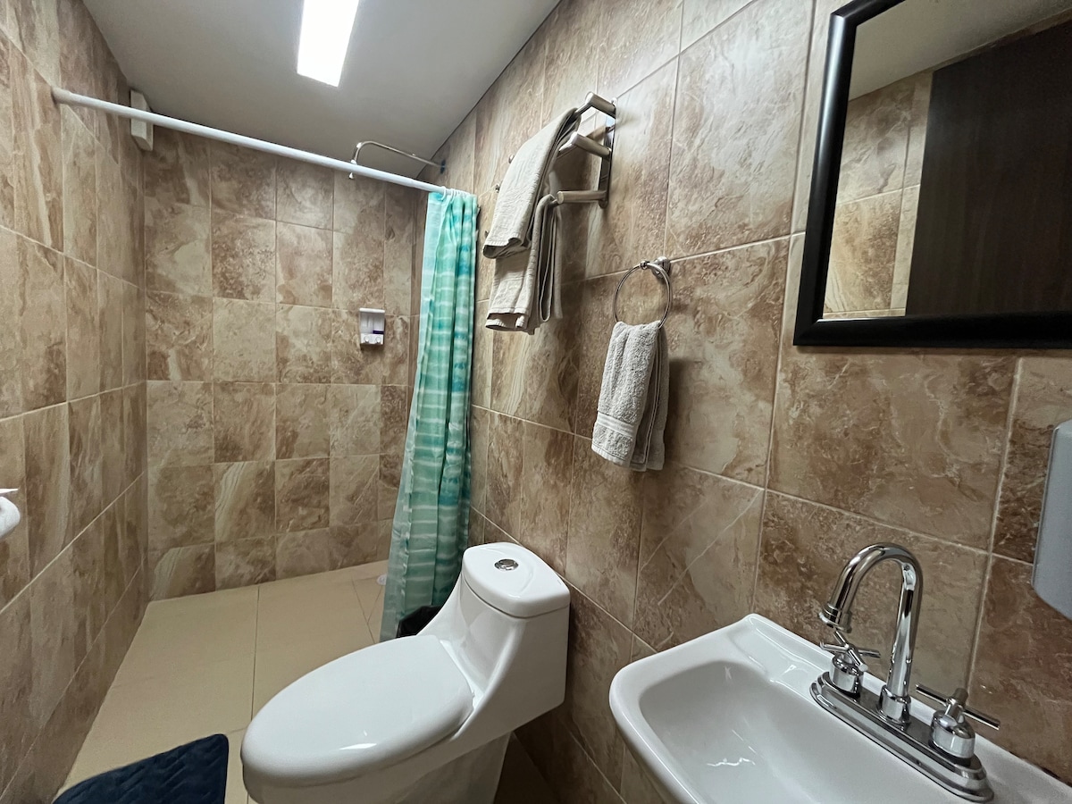 The bathroom features beige tiled walls providing a neutral backdrop. A modern toilet is positioned next to a white sink with a sleek faucet. An azure towel lies on the floor, while towels hang on a rack and hooks nearby. Bright lighting illuminates the space.