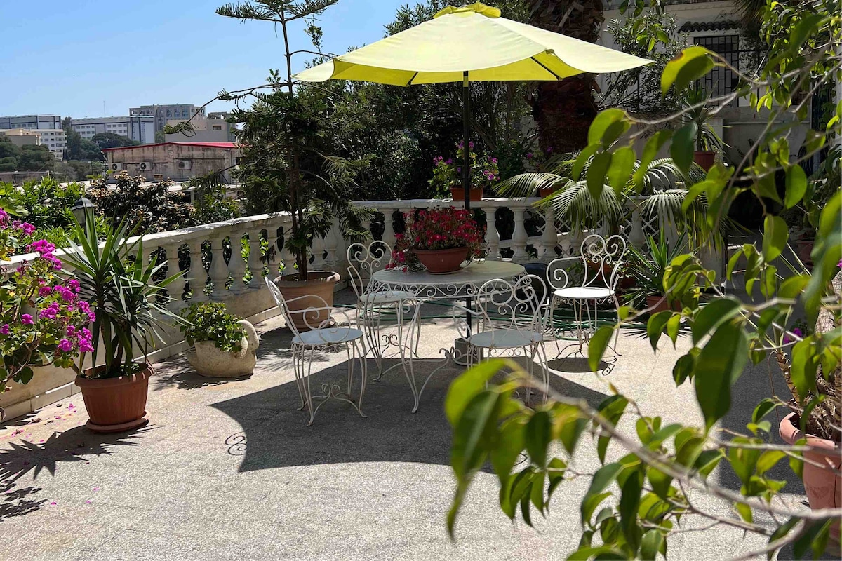A spacious terrace is surrounded by greenery and adorned with potted plants and flowers. A round table with four chairs is shaded by a large umbrella. The serene space features a decorative railing, offering a calm atmosphere for relaxation.