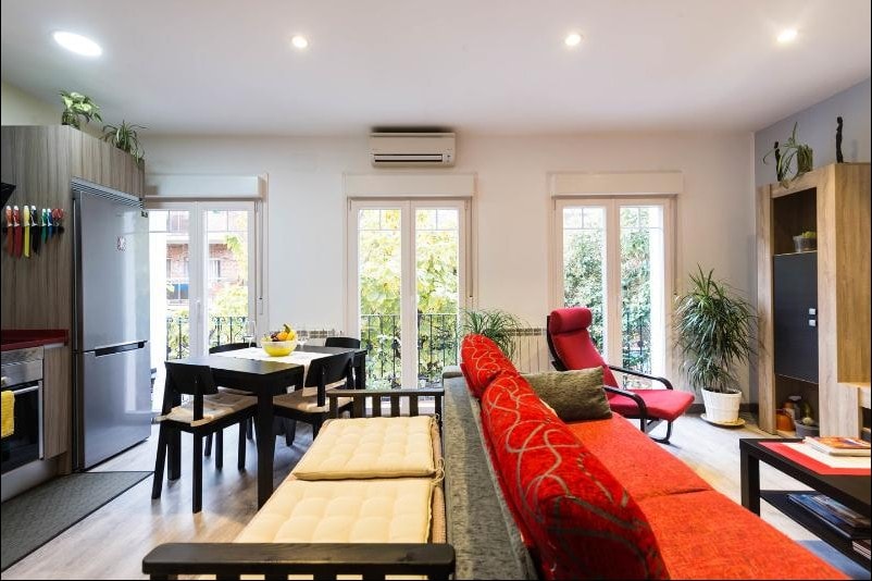 The living area features a red sofa bed facing a table set for four. Large windows with white frames let in natural light and provide views of greenery outside. A compact kitchen with modern appliances is visible, along with plants positioned around the space.