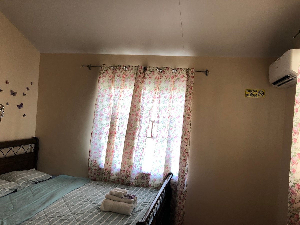 A cozy bedroom features a double bed with a light blue and white patterned bedding set, complemented by floral curtains allowing natural light to filter through. An air conditioning unit is mounted near the ceiling, and a set of towels is neatly arranged on the bed.