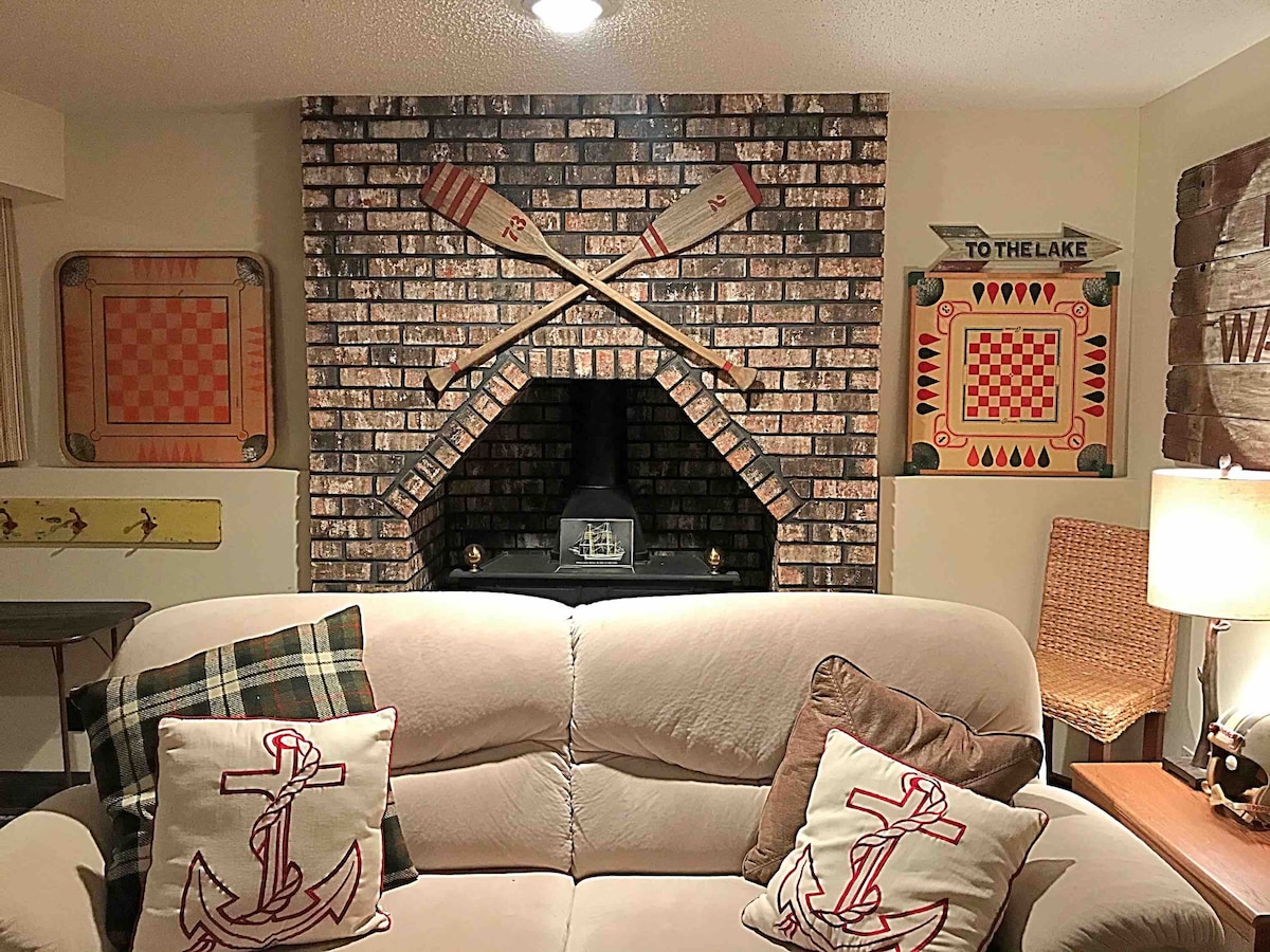 A brick fireplace is centered on the wall, adorned with two crossed oars. On either side, decorative artwork featuring checkered patterns and wooden signs contributes to a cozy ambiance. A light-colored sofa with anchor-themed cushions provides comfortable seating in the space.