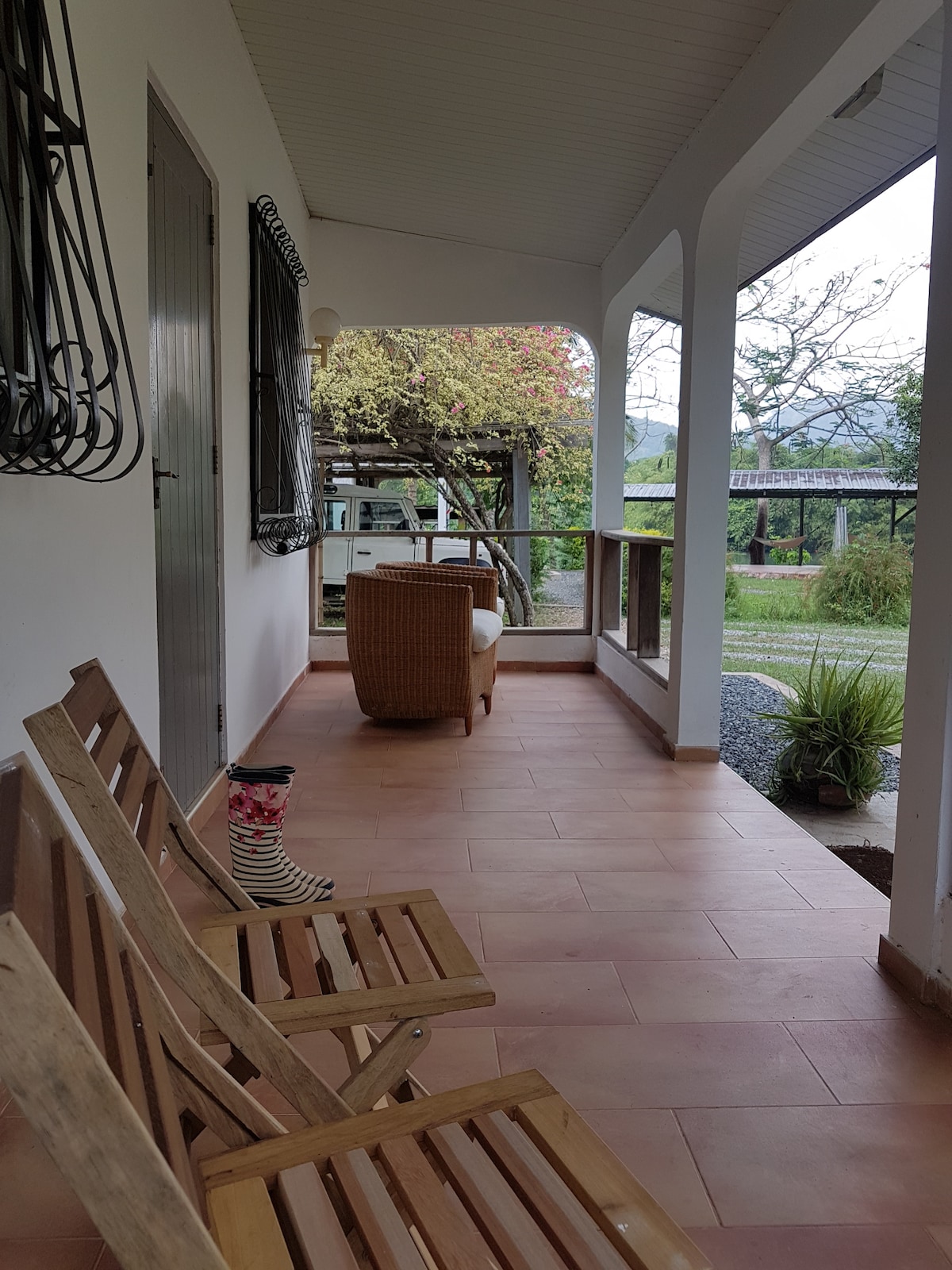 A private veranda is furnished with wooden chairs and a cozy rattan seating area. Lush greenery is visible through the open space, inviting relaxation. The floor features terracotta tiles, while elegant structures and plants provide a tranquil ambiance.