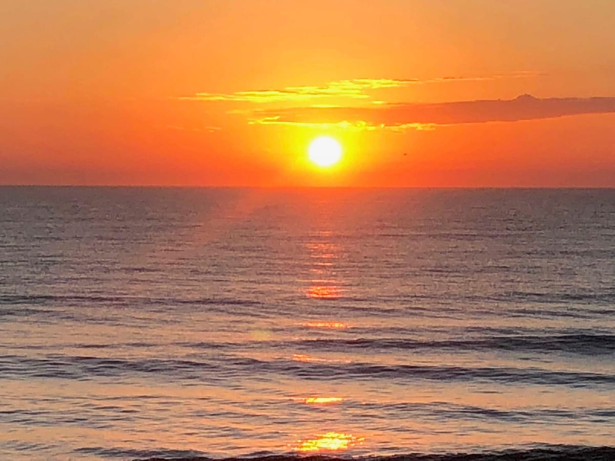 The sun is seen rising over a calm ocean, casting warm orange and yellow hues across the sky and reflecting on the water's surface. Gentle waves are visible, creating a serene coastal scene.