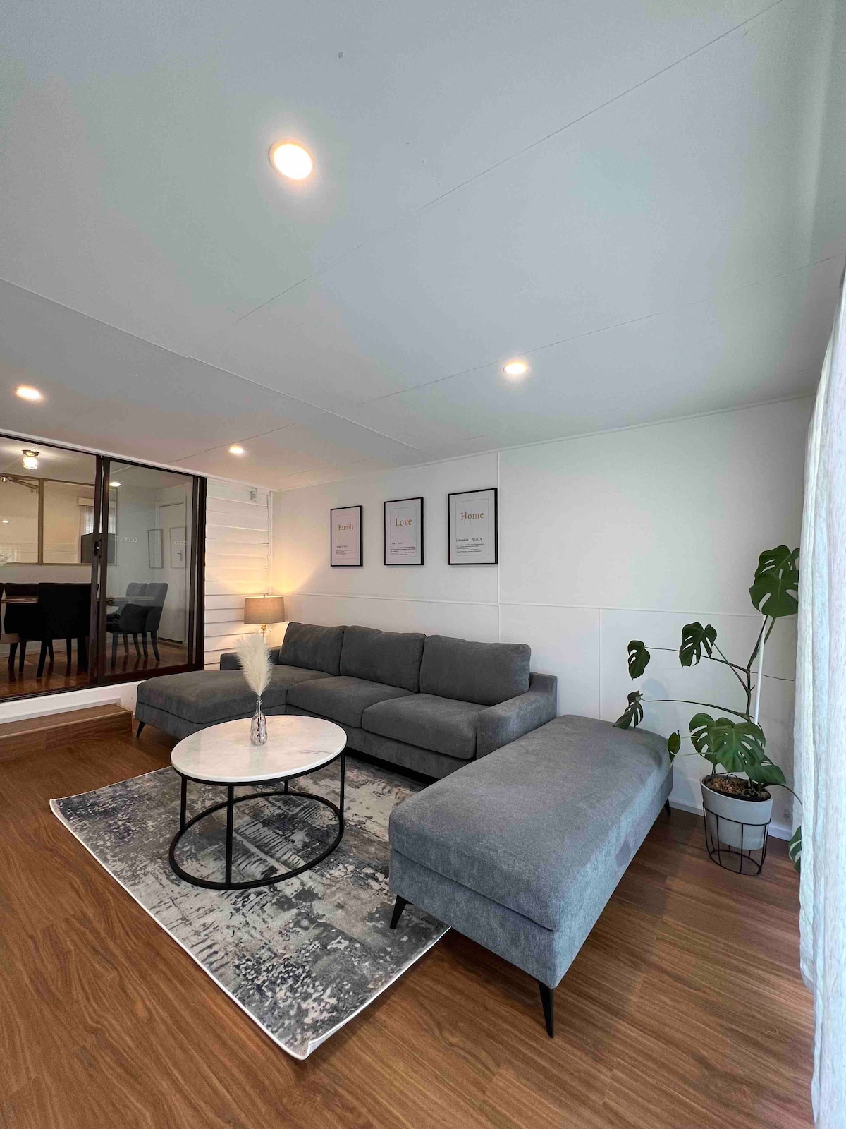 A spacious living room features a comfortable gray sofa with an accompanying chaise lounge. A round coffee table sits in front of it, placed on a patterned area rug. Natural light enters through large glass doors leading to the dining area, enhancing the inviting ambiance.