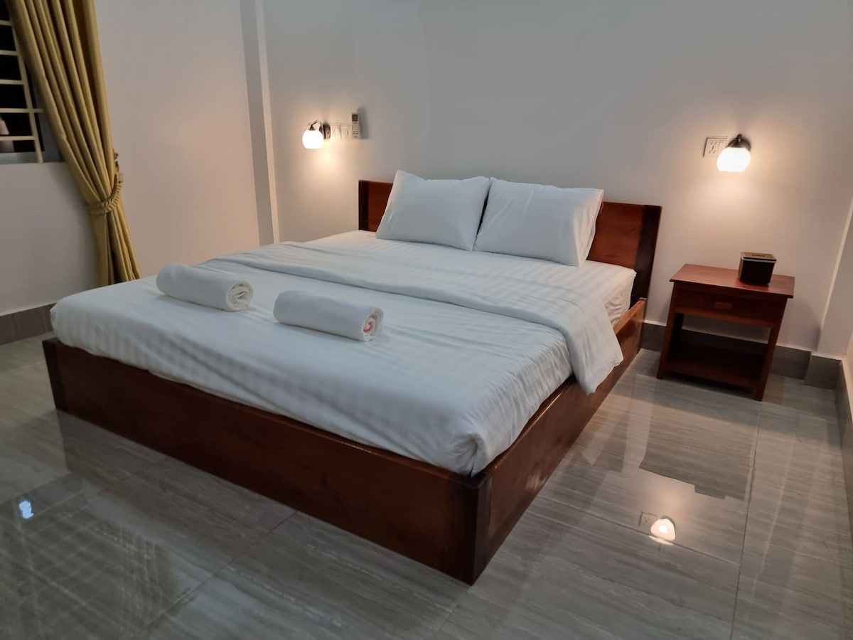 A spacious sleeping area features a large wooden bed with a light-colored striped duvet. Two rolled towels are neatly placed on the bed. A bedside table with a small lamp is positioned next to the bed, while soft lighting enhances the calm atmosphere.