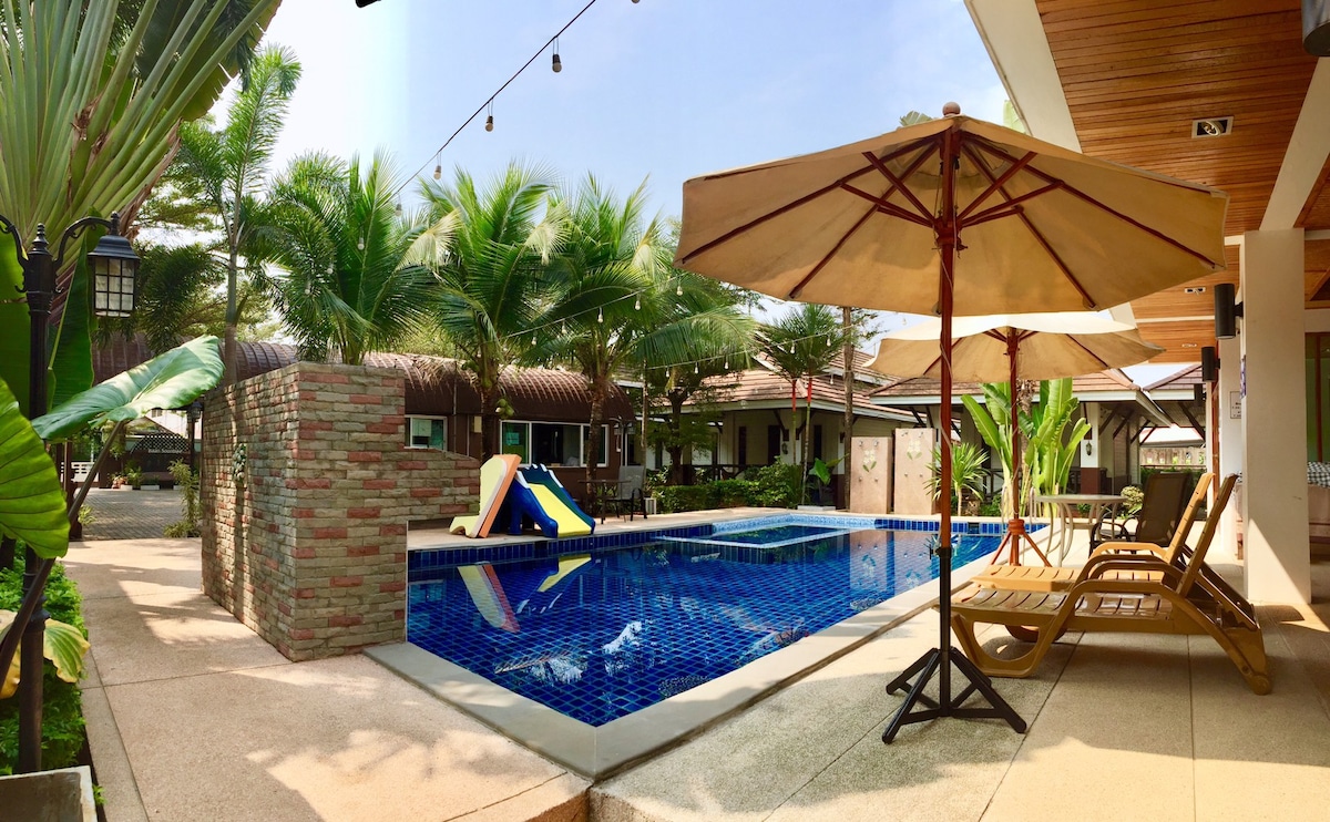A communal swimming pool is surrounded by a sun deck featuring lounge chairs and umbrellas. Tropical plants add greenery to the area, while a waterslide provides a playful element. The architecture in the background includes multiple houses, creating a serene and inviting outdoor environment.