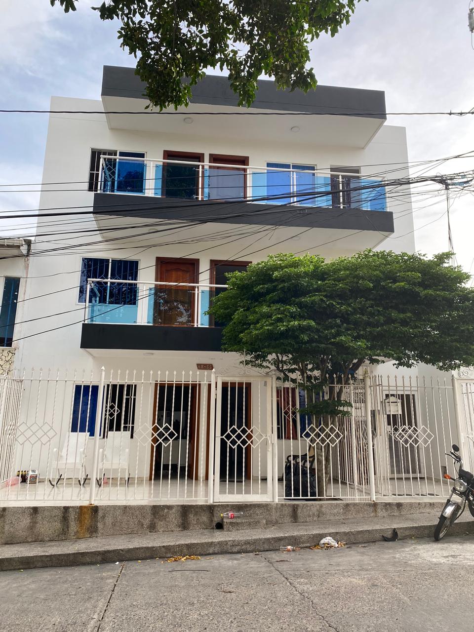 A modern three-story apartment building features a striking white exterior with black accents. Multiple balconies display glass railings, while a decorative fence encloses the entrance. Lush green trees are situated in front, enhancing the building’s appeal and creating a welcoming environment.