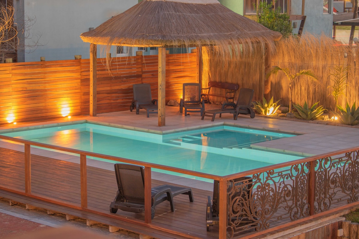 A sun-soaked outdoor space features a rectangular swimming pool surrounded by a wooden deck. Lounge chairs are arranged for relaxation beneath a thatched roof structure. Soft lighting enhances the area, creating an inviting atmosphere for leisure and gathering.