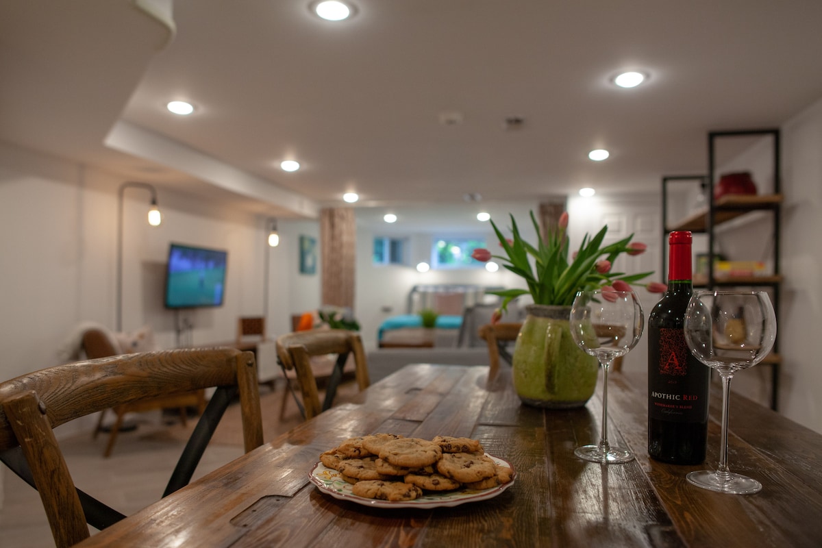 A welcoming dining area is presented with a wooden table featuring a plate of cookies and a bottle of red wine, accompanied by two glasses. In the background, a cozy seating area is visible with a television, and vibrant flowers are displayed in a green vase.