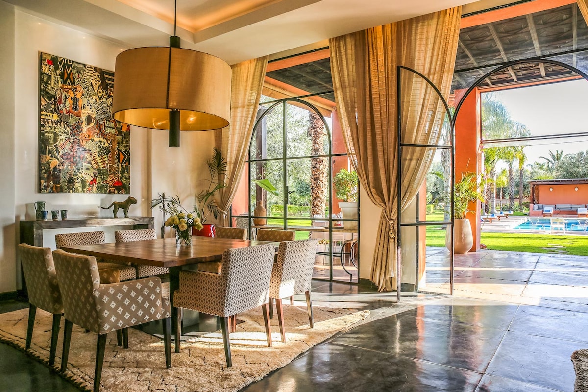 A dining area features a wooden table surrounded by patterned chairs, illuminated by a large pendant light. Floor-to-ceiling arched windows offer views of the garden and pool, enhancing the open layout. A decorative wall art piece adds a vibrant touch to the space.