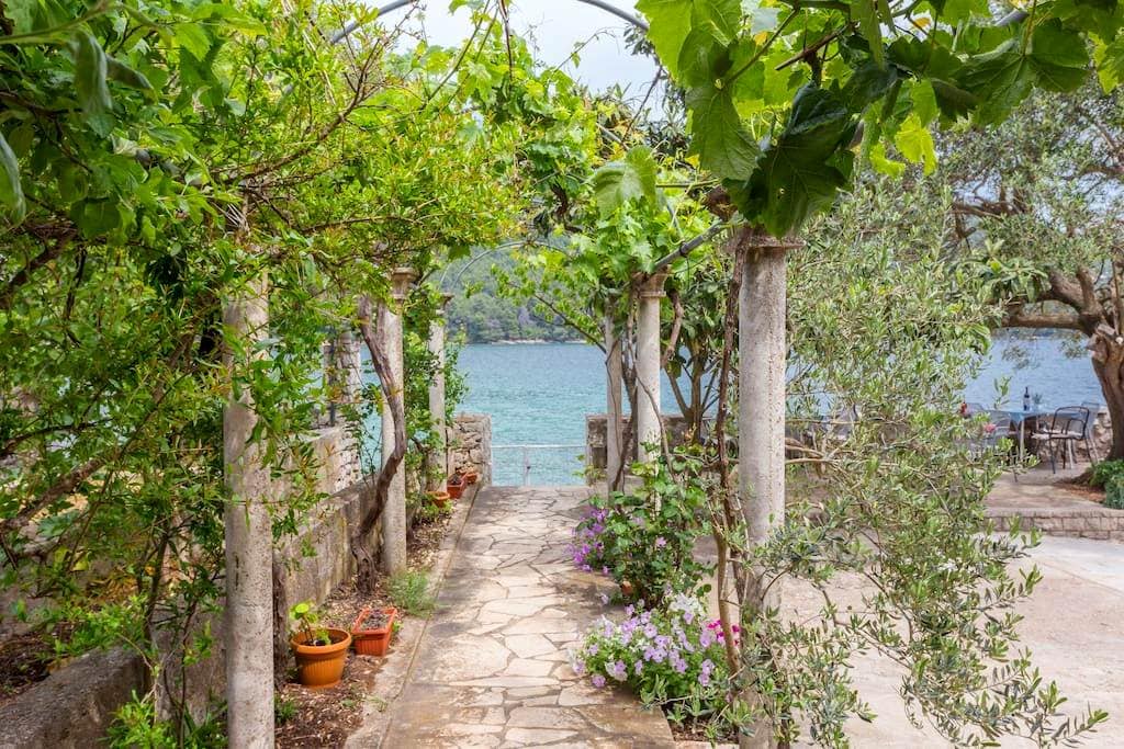 A stone pathway is framed by lush greenery and flowering plants, leading towards the water's edge. Columns support the overhanging vines, creating a shaded walkway that guides the eye to the tranquil sea visible in the background.