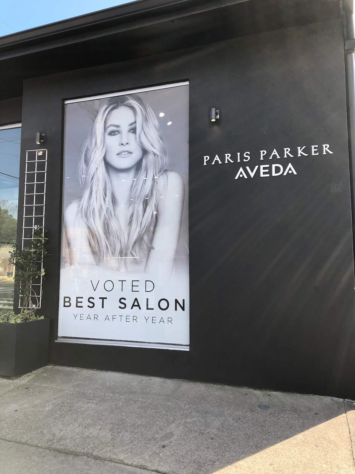 The exterior of a salon is featured, with a prominent advertisement displayed. The black facade is complemented by a large window poster showcasing a model with long hair. The salon's name, 'Paris Parker Aveda,' is visible in elegant lettering beside the entrance.