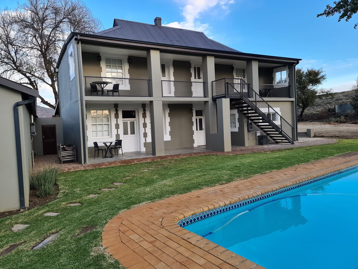 A two-story building is shown with a modern design, featuring a balcony on the upper level and outdoor seating arrangements. A well-maintained swimming pool is visible in the foreground, surrounded by a neat pathway and green grass, adding to the outdoor appeal.