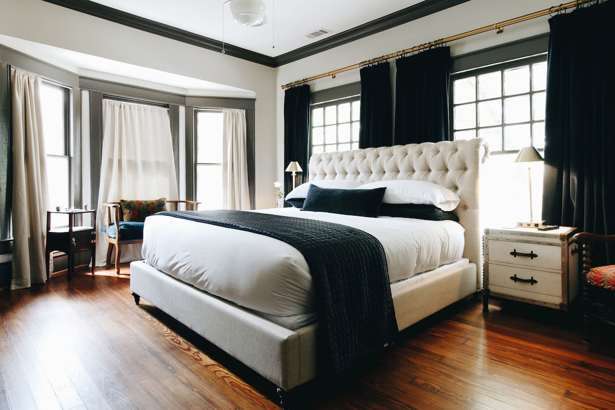 A spacious bedroom features a king size bed with plush white bedding and a textured dark throw. Two large windows allow natural light to fill the room, accented by elegant dark curtains. Cozy chairs are positioned at the side, creating inviting seating areas.
