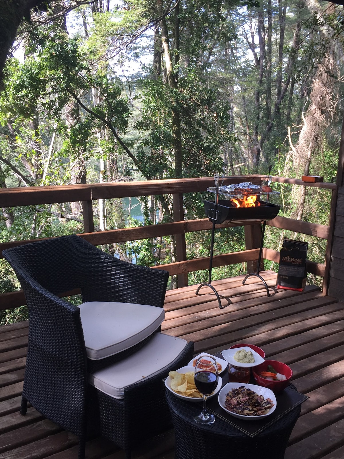 A wooden deck surrounded by lush greenery features a grill with flames, accompanied by two black wicker chairs with light cushions. A table displays a spread of food, including appetizers and main dishes, inviting guests to enjoy outdoor dining in a natural setting.