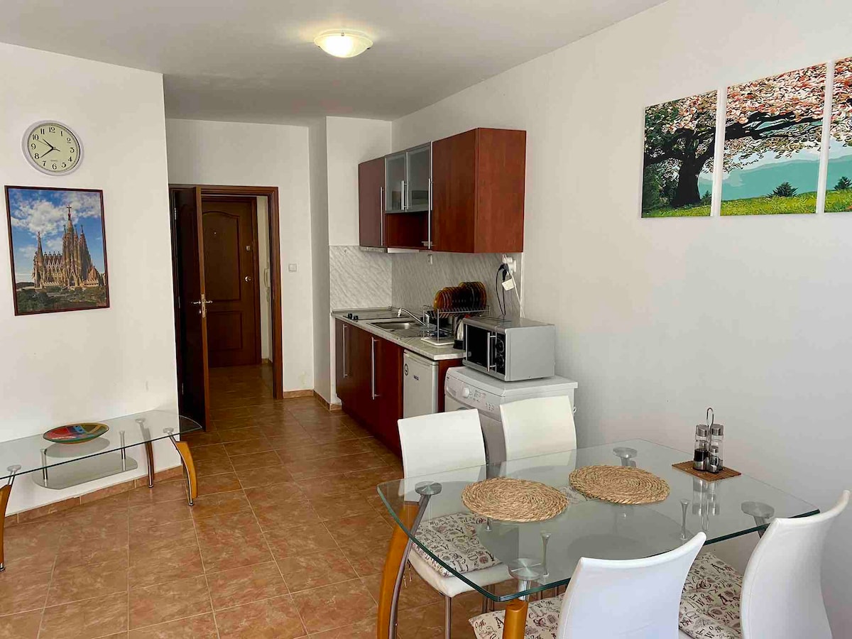 A modern kitchen area is visible, featuring wooden cabinets and essential appliances like a microwave and stove. A dining table with white chairs is set for meals. Wall art depicting nature adds a touch of color, while a clock provides a practical element.