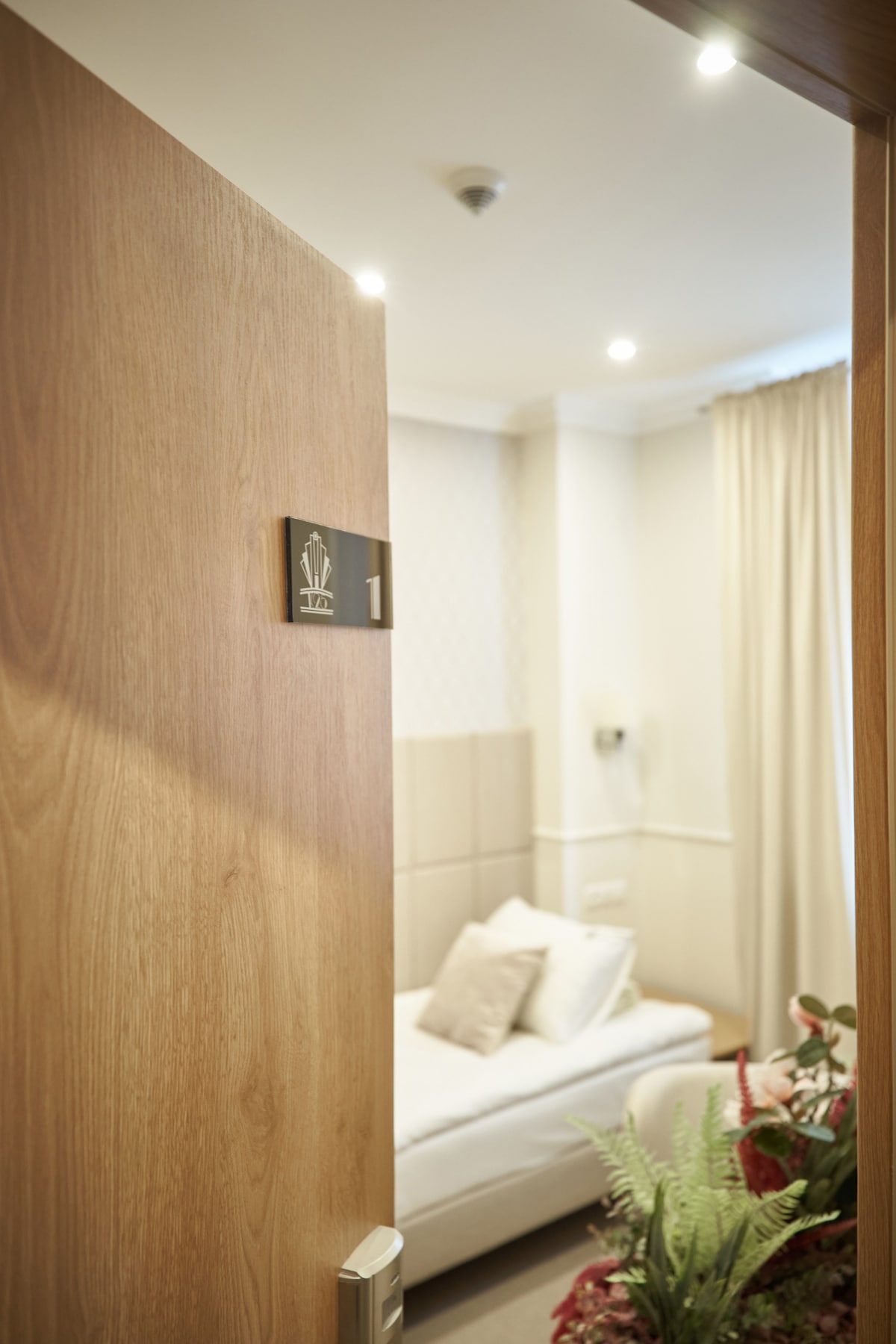 A doorway opens to reveal a modern room featuring a single bed with neutral linens. Soft lighting creates a calming ambiance, and sheer curtains frame the window, allowing natural light to fill the space. Decorative plants add a touch of greenery.