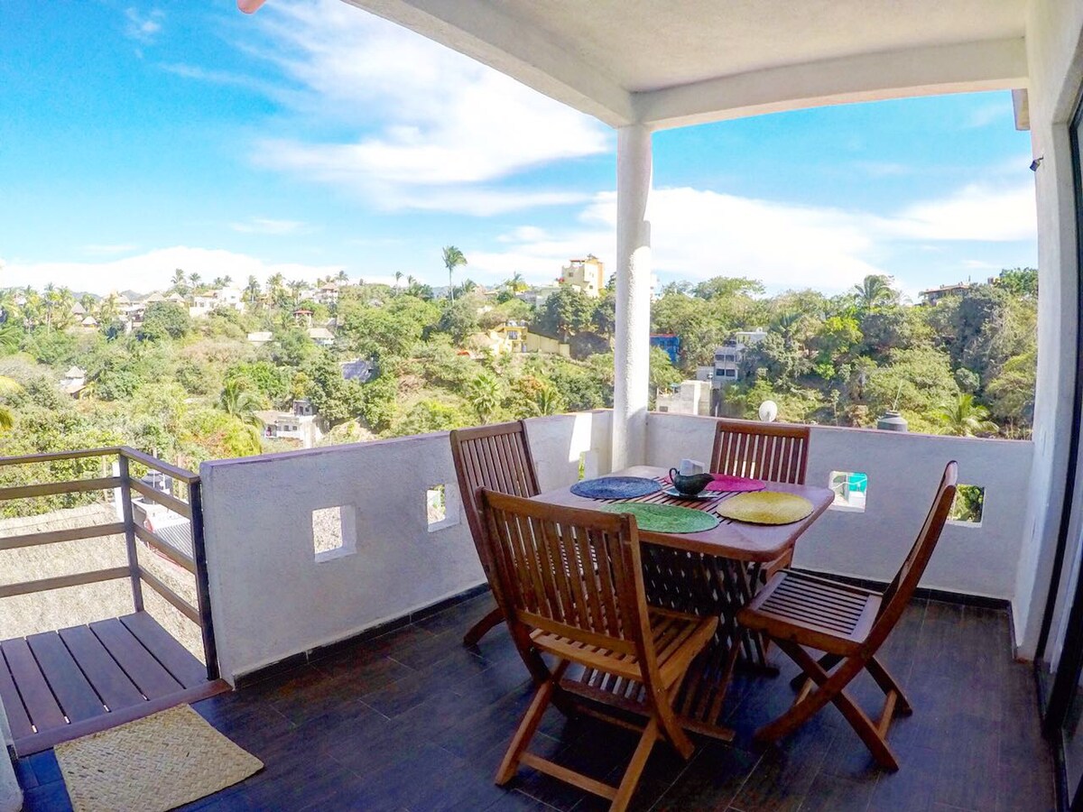 A spacious terrace features a wooden dining table surrounded by matching chairs, offering beautiful views of the lush surroundings. Colorful placemats and a decorative bowl are placed on the table, while the sky above is bright and clear, indicating pleasant weather.