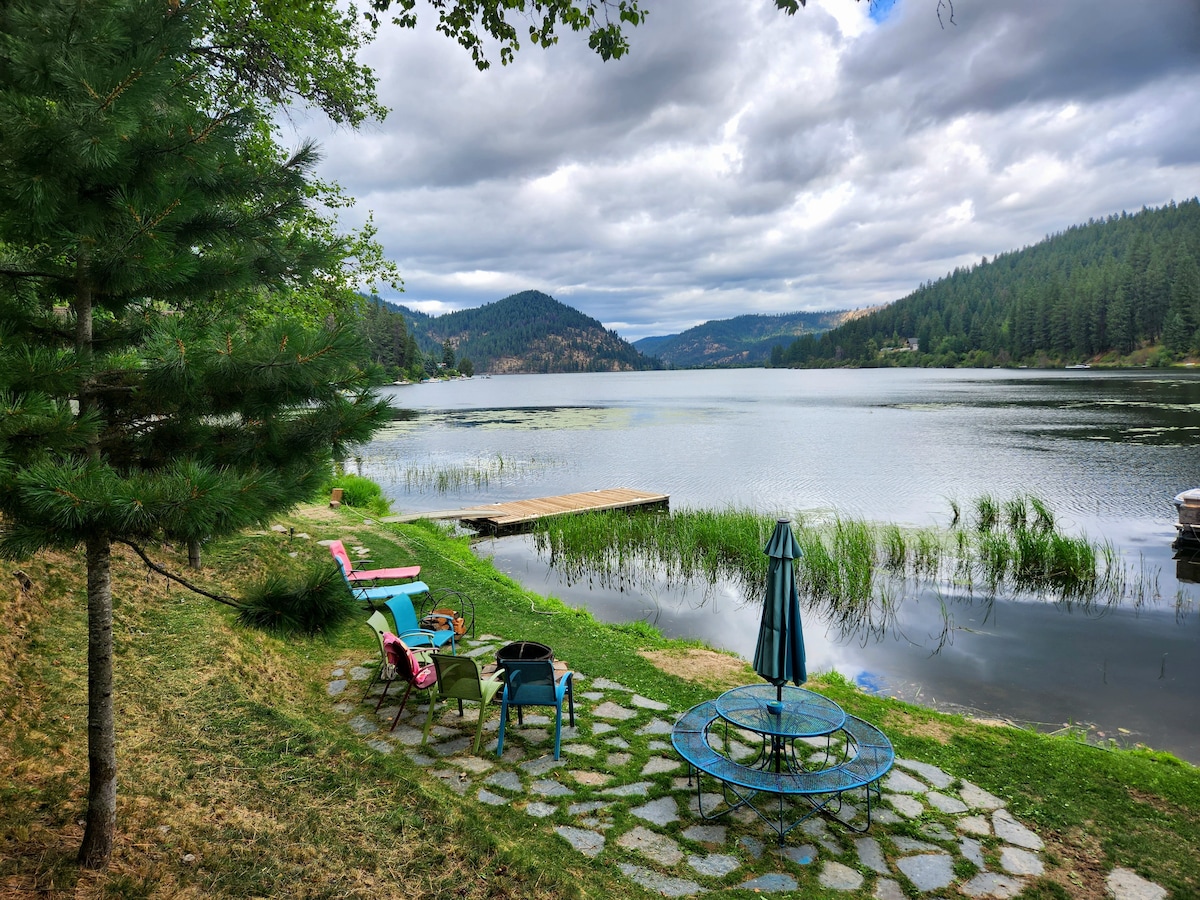 A serene lakefront view showcases a small dock extending into calm waters. Colorful chairs are arranged around a circular table under an umbrella. Lush greenery and distant mountains frame the scene, with water lilies and aquatic plants gently swaying in the breeze.
