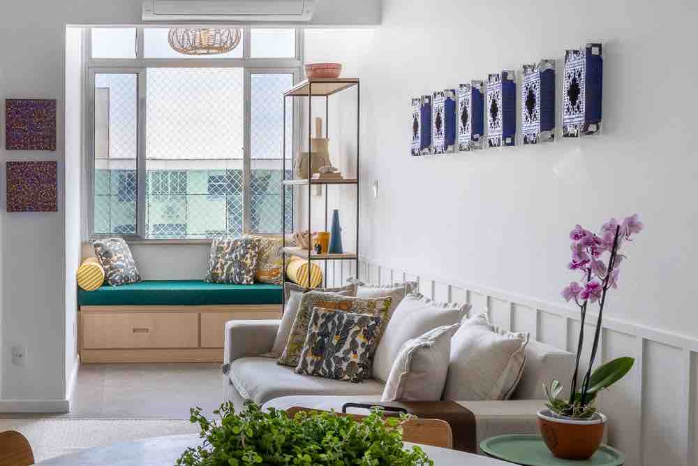 A well-lit living area features a cozy seating nook with cushions, accompanied by decorative wall art. The space is complemented by a modern sofa adorned with patterned pillows and a round dining table topped with greenery and a vibrant orchid.