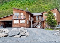The exterior of a wooden lodge is depicted, showcasing a two-story structure. A central staircase leads to a second-floor entrance, surrounded by balconies. Large windows allow natural light, with a gravel driveway and boulders in the foreground. Green hills rise in the background.