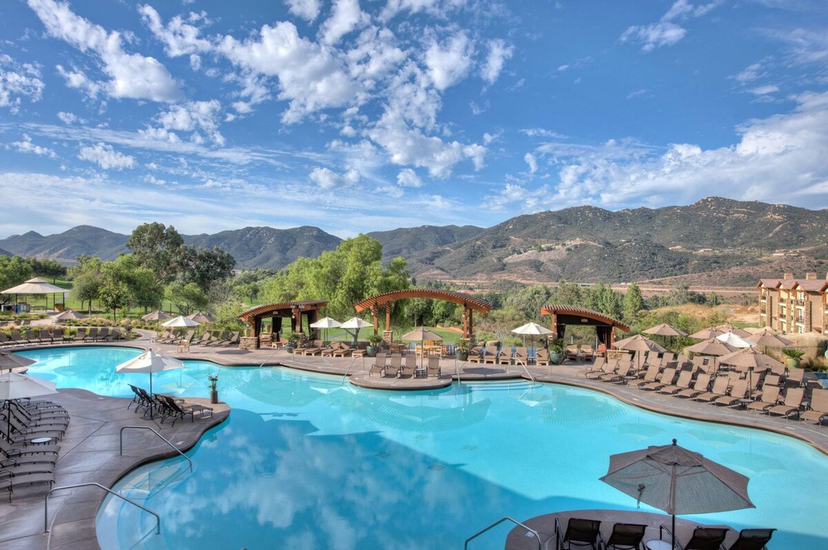 A large swimming pool is visible, surrounded by lounge chairs and shaded umbrellas. The area features elegant wooden structures providing shade, with lush greenery and rolling hills in the background under a partly cloudy sky.