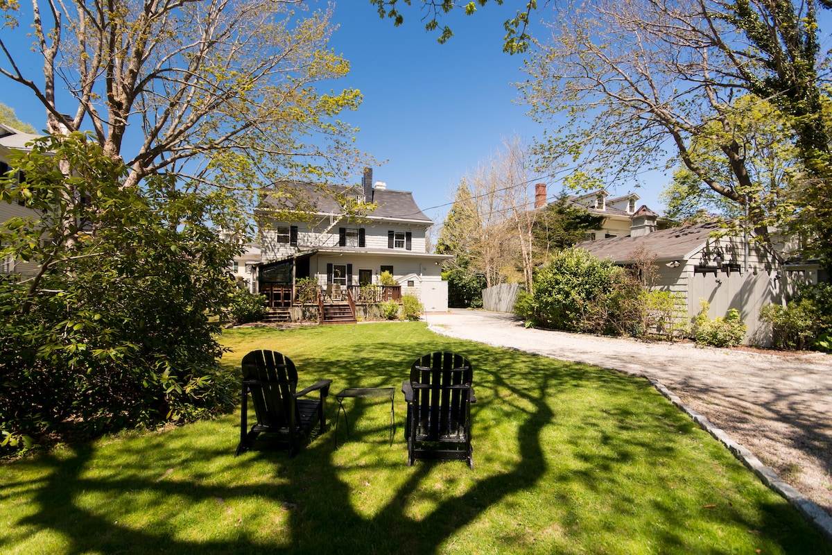 A spacious outdoor area is featured, showcasing a well-maintained lawn with two black Adirondack chairs positioned on a grassy patch. Surrounding trees provide shade, with the inn's structure visible in the background, creating a peaceful and inviting environment.