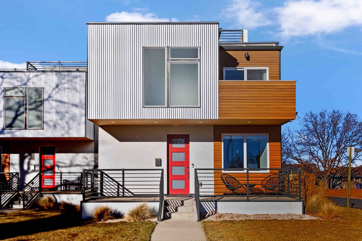 A modern three-story home is presented, featuring a striking facade with a combination of vertical and horizontal wooden paneling. A vibrant red front door stands out against the neutral exterior. Adjacent are two minimalist chairs positioned on a small patio area, with clear skies overhead.