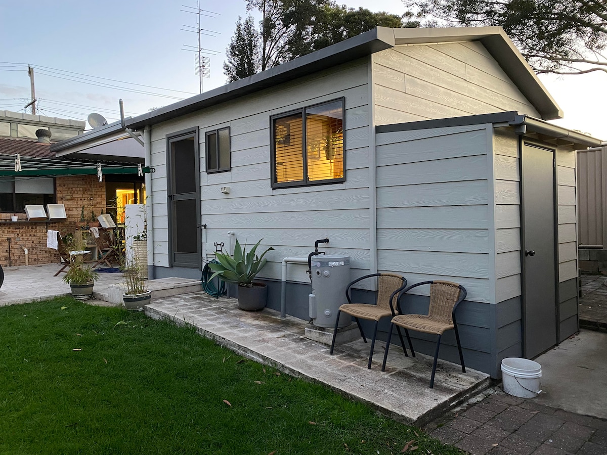 The exterior of the Pallet Cabin is shown, featuring a light grey and dark grey color scheme. A small porch area is highlighted with two outdoor chairs and a potted plant. The entrance is framed by a window that emits warm light, surrounded by a grassy yard.
