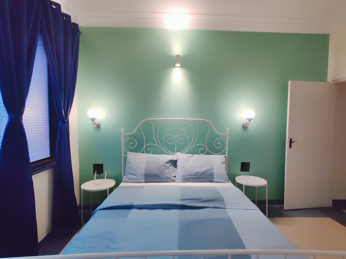 A spacious bedroom is presented with a white metal bed topped with light blue bedding. Two bedside tables with lamps flank the bed, while a deep blue curtain frames the window, allowing soft light to enter. A light green wall enhances the room's fresh ambiance.