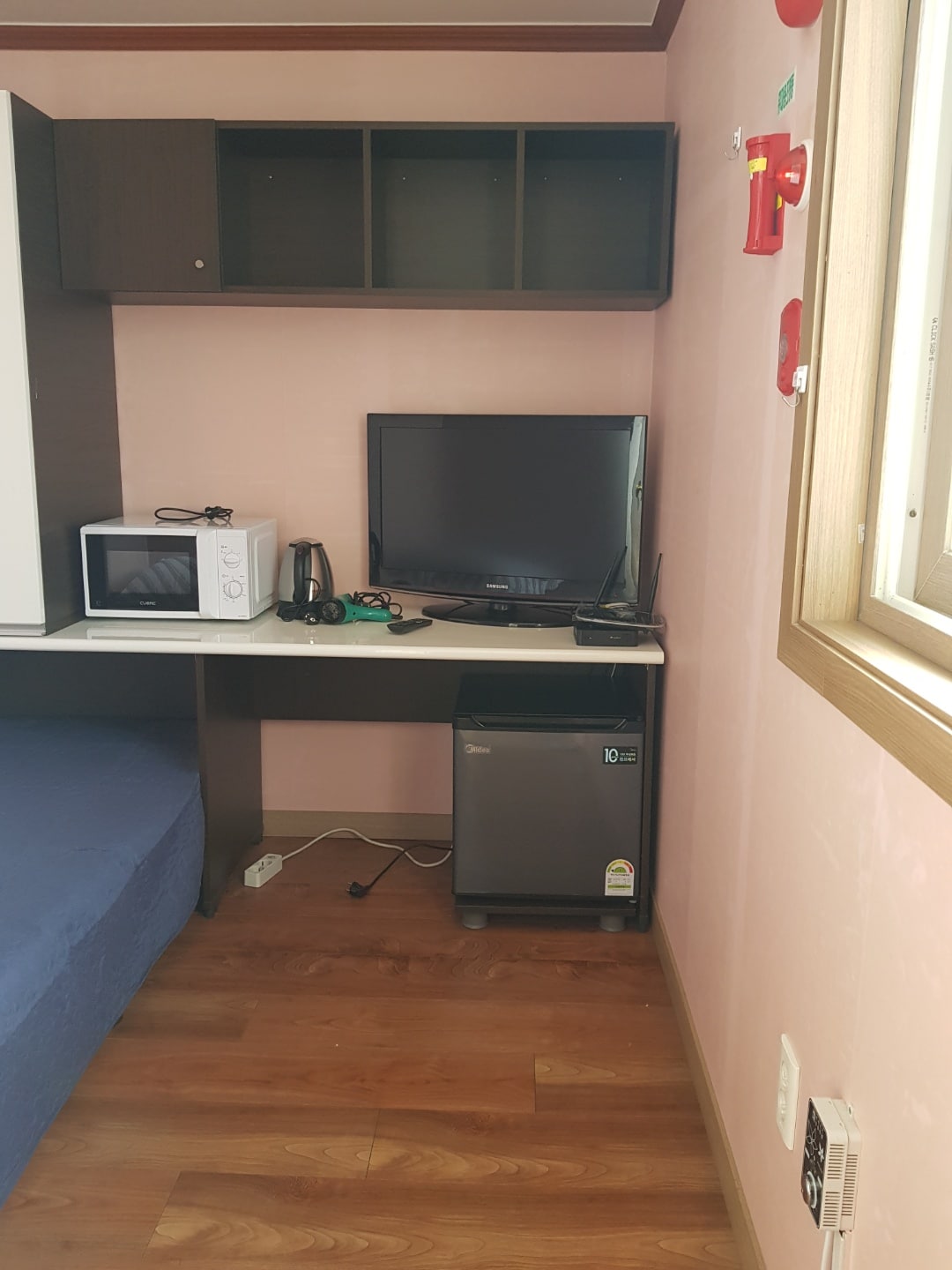 A compact workspace is featured, with a desk holding a television, microwave, and kettle. A small refrigerator is positioned below the desk. Light-colored walls enhance the brightness of the room, while a window allows natural light to enter.