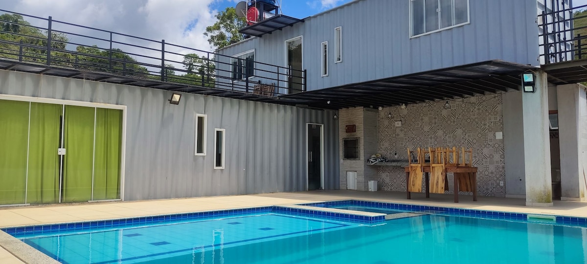 A modern two-story container home is depicted beside a clear swimming pool. The pool is surrounded by a spacious deck, featuring a covered barbecue area with a wood table and chairs. Large windows provide ample light, complemented by a green sliding door.