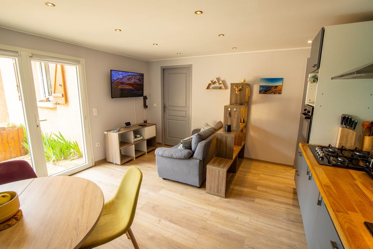 An open living area features a cozy sofa positioned near a television console. Bright, natural light floods through double glass doors, illuminating the wooden dining table with colorful chairs. Soft gray walls and light wood flooring create a calm ambiance throughout the space.