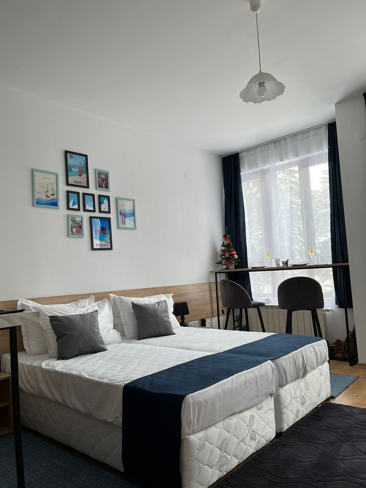 A bright sleeping area is presented with a spacious bed adorned with a navy blue bedspread. A set of decorative photographs hangs on the wall above, while a small bar table with two stools offers additional seating near the large window dressed with sheer curtains.
