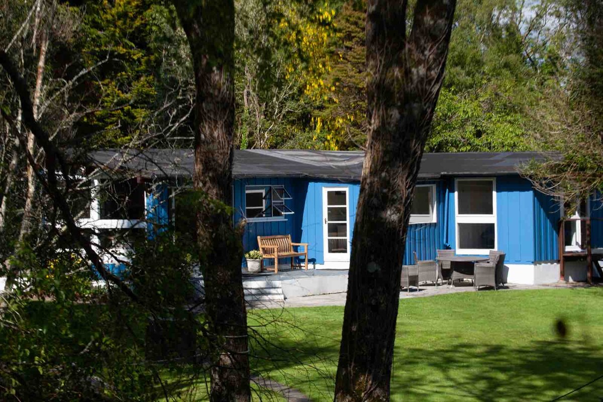 A timber cottage features a blue exterior, surrounded by lush greenery. A shaded porch area holds a wooden bench and outdoor seating. The spacious lawn showcases well-maintained grass with mature trees providing a natural backdrop.