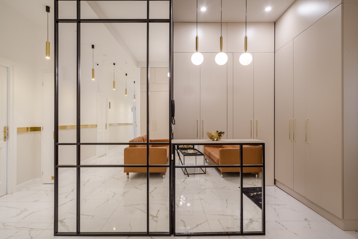 A modern entryway features a sleek glass partition and neutral-toned walls. A comfortable seating area with a tan couch and a coffee table is visible, while pendant lights illuminate the space. Marble flooring reflects the soft lighting, creating an inviting atmosphere.