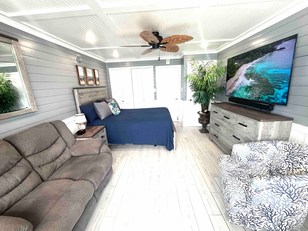 The spacious oceanfront room features a queen-sized bed covered in deep blue bedding. A large flat-screen TV is mounted on the wall opposite a comfortable seating area with a recliner and a patterned accent chair. Natural light floods the room through sliding glass doors leading to a private balcony.