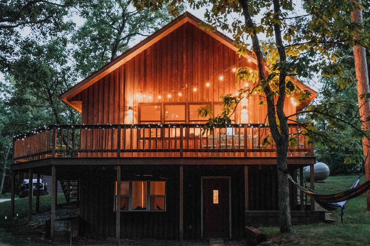 A chalet-style home features warm ambient lighting and a spacious deck surrounded by trees. The upper level showcases large windows, providing a view of the outdoors. A hammock is visible nearby, inviting relaxation in the natural setting.