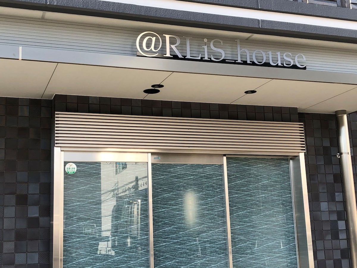The entrance features a sleek facade with a contemporary design. A large glass door is complemented by a stylish sign that reads '@RLis_house', showcasing a modern aesthetic. The exterior walls are adorned with a textured pattern, enhancing the overall appearance.