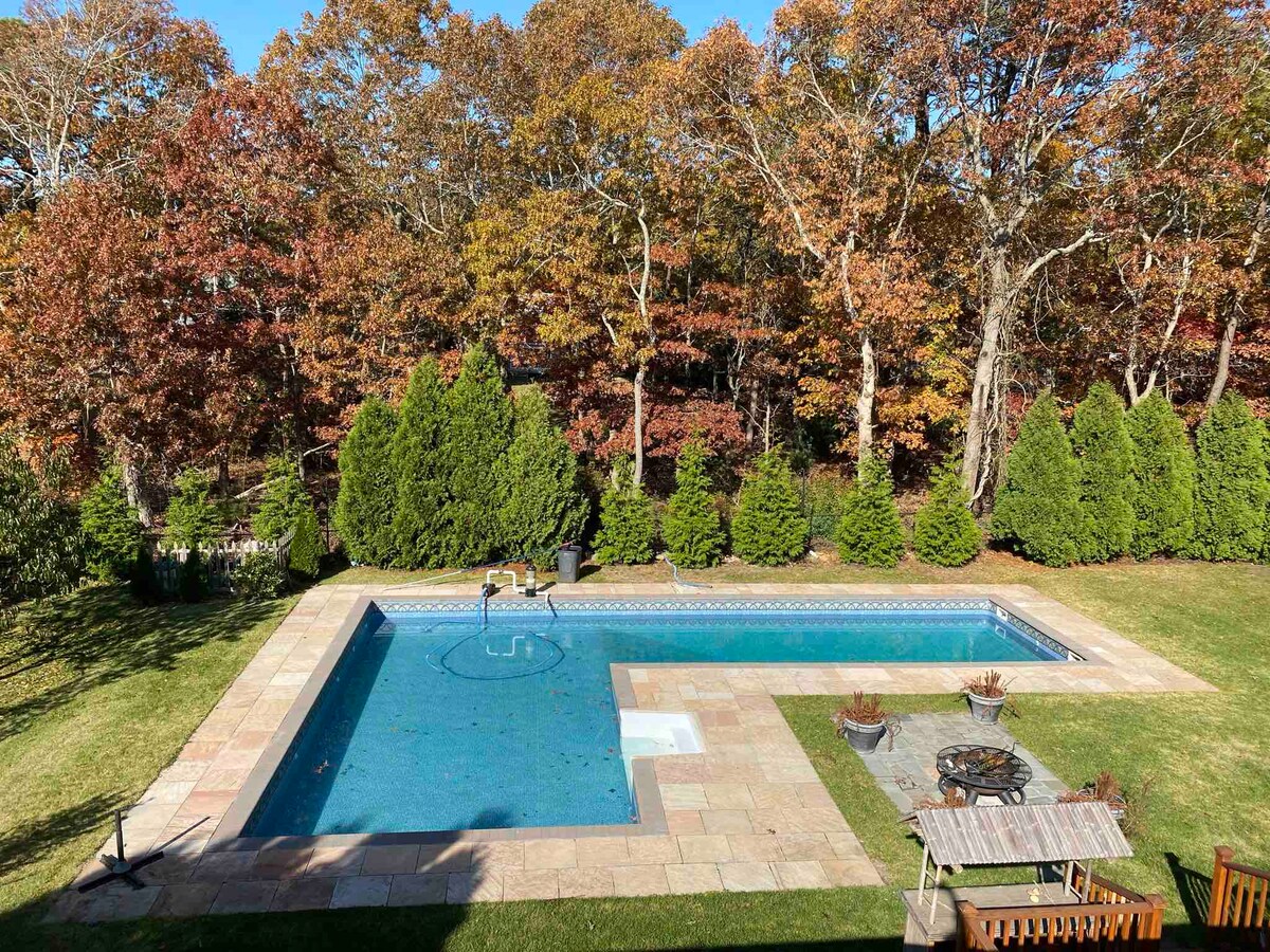 An outdoor area features a heated saltwater pool surrounded by a spacious patio. Lush greenery and trees provide privacy, while an outdoor seating arrangement offers a place to relax. The setting showcases seasonal foliage, creating a serene and inviting atmosphere.