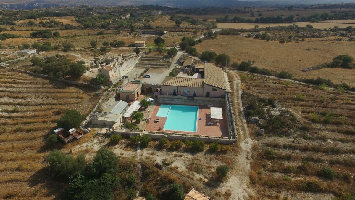 An aerial view showcases the agriturismo nestled among rolling hills, surrounded by cultivated fields. A large outdoor pool is centered within the property, complemented by sun loungers and shaded areas. The rustic buildings exhibit a traditional architectural style, set against a backdrop of natural scenery.