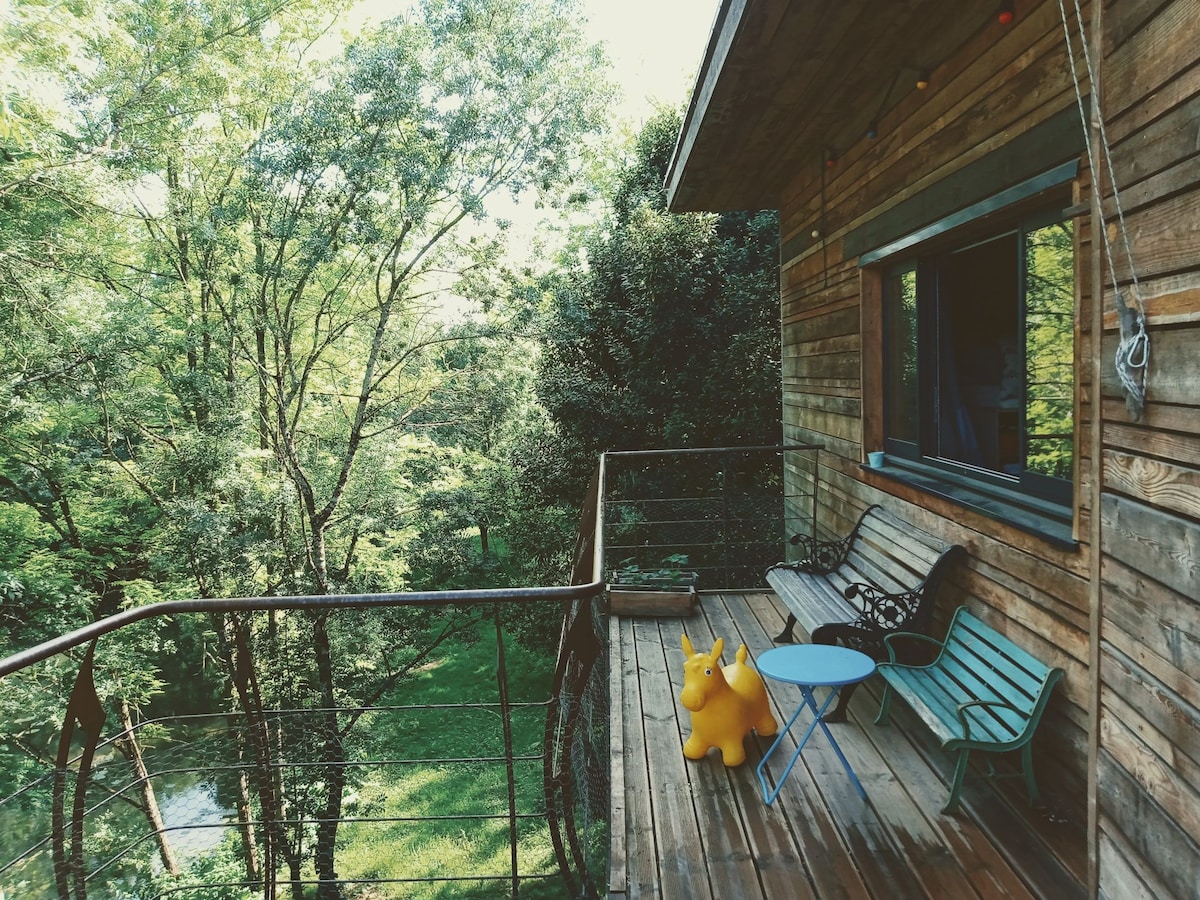 A wooden terrace is equipped with a small blue table and assorted seating, including a bright yellow sculpture that adds a playful touch. The surrounding lush greenery offers a serene view, with trees filtering soft sunlight into the space.