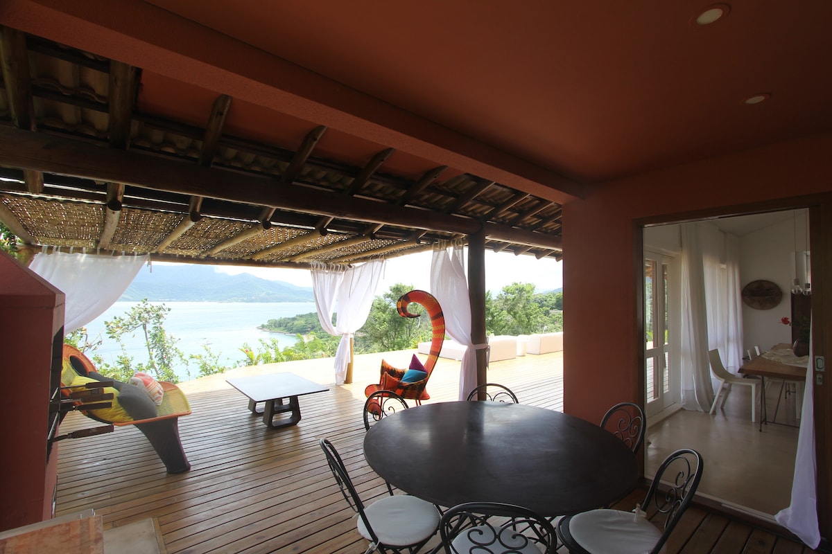 An outdoor terrace features a wooden deck with a round dining table surrounded by black chairs. A lounge chair with a colorful design and a hammock are positioned nearby, offering views of the sea through sheer white curtains that gently frame the space.