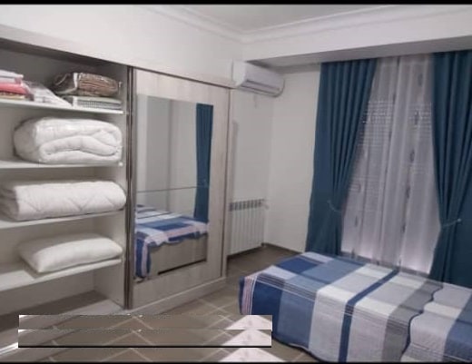 A comfortable bedroom features a spacious bed covered in a patterned blanket. A mirrored wardrobe provides storage, alongside neatly stacked linens on open shelves. Blue curtains frame a window, allowing natural light to fill the room, while a radiator offers warmth.