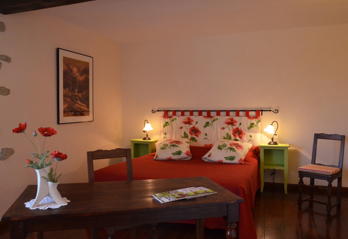 A cozy bedroom is highlighted by a spacious bed adorned with floral-patterned pillows against a decorative headboard. Two lamps illuminate the nightstands, while a wooden table with a flower vase adds to the charm. A piece of art decorates the wall, enhancing the overall ambiance.