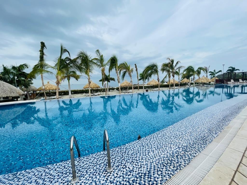 A large swimming pool features a mosaic tile border, reflecting the surrounding palm trees. Beach huts can be seen in the background under a cloudy sky, enhancing the tranquil atmosphere. Lounge chairs line the pool area, providing spots for relaxation and sunbathing.