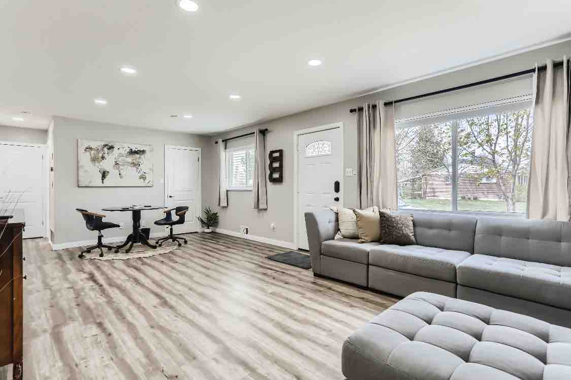 An open-plan living room features a large sectional sofa and a chic work area with two chairs. Natural light enters through the large windows, and a world map hangs on the wall. Gleaming hardwood floors complement the space, creating a bright and functional atmosphere.