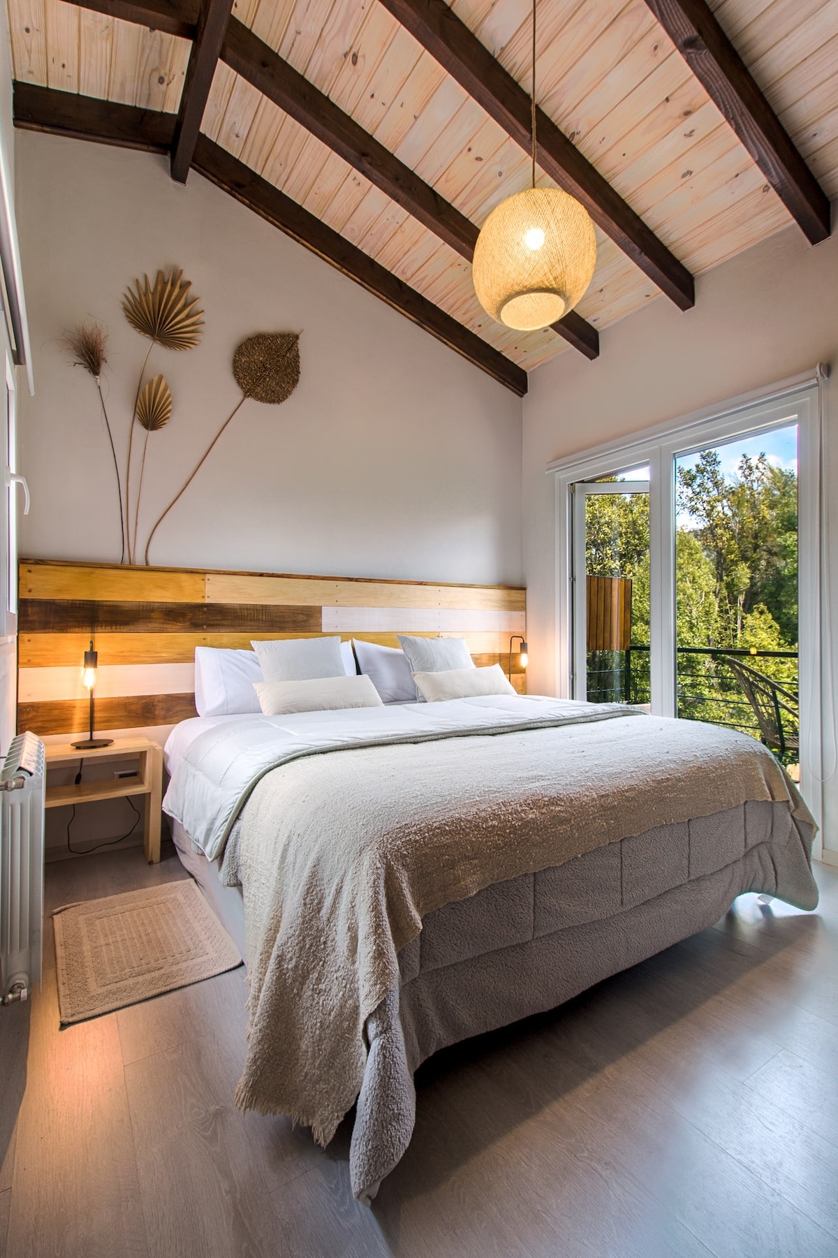 A serene bedroom features a spacious bed with a light-colored blanket and decorative pillows. Natural light enters through large glass doors that lead to a balcony. Wooden accents on the wall and a modern lighting fixture contribute to a calming atmosphere.