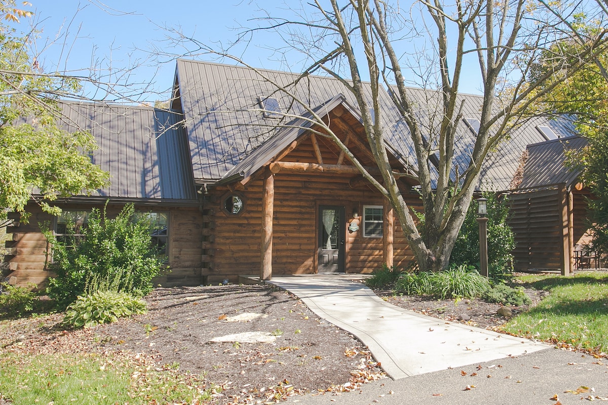 The exterior of the cabin features a rustic log construction with a pitched roof. A paved walkway leads to the entrance, surrounded by well-maintained landscaping, including bushes and a mature tree. Natural light illuminates the façade, enhancing its welcoming appearance.