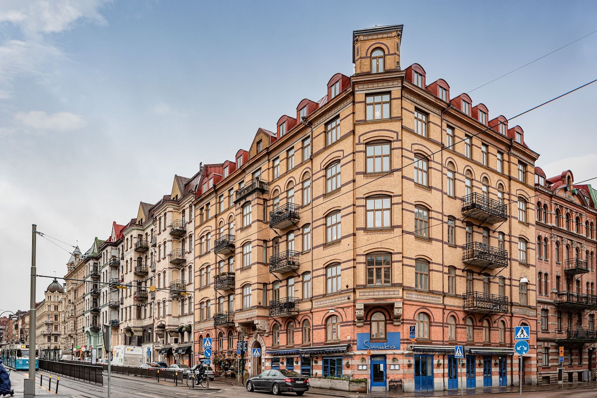 A vintage-style building is depicted with a mix of architectural details. The façade features red tiles and numerous windows, while decorative balconies add character. The street below shows clear signage and a tram line, indicating a central urban location.